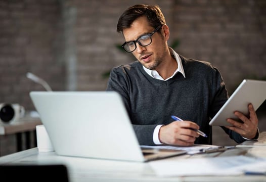 Un hombre sentado en su sitio de trabajo, revisando información en su computador portátil y tablet.